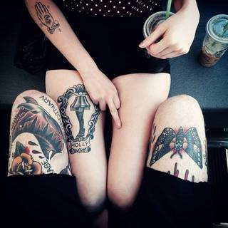 Dziewczyna z tatuażem na nodze i chłopak zakrywający jej nogi swoimi nogami, na których są dwa tatuaże. ><