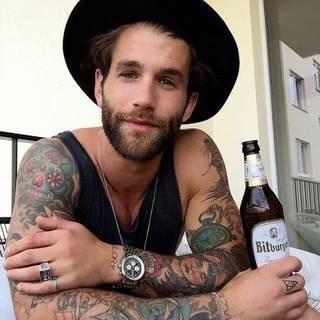 Mężczyzna z tatuażami na rękach (np. zegar, postać kobiety, napis) oraz pojedynczym tatuażem na palcu.